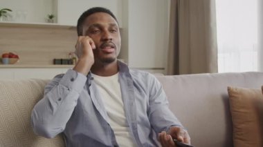 Man using TV remote. Young adult talking on a phone in his apartment