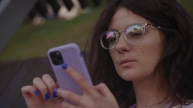 Gözlüklü kıvırcık kadın akıllı telefondan bakıyor. Parkta internette sörf yapan beyaz bir kız öğrencinin fotoğrafını çek.