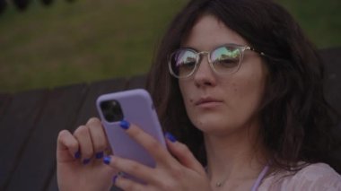 Curly woman in glasses browsing on smartphone outside. Close up of a caucasian female student online surfing in the park