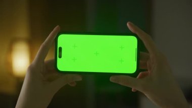 Horizontal smartphone with green screen close up. Caucasian woman hands holding a smart phone with chroma key indoors slow optical zoom in