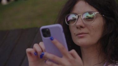 Curly woman in glasses browsing on smartphone in park. Close up of a caucasian female student online surfing