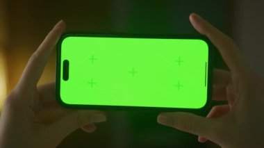 Horizontal smartphone with green screen close up. Caucasian woman hands holding a smart phone with chroma key indoors slow optical zoom out