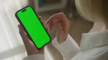 Tanımlanamayan bir kadın çift tıklıyor ya da omzunun üstünden yeşil ekran akıllı telefona tıklıyor. Beyaz kadın 60 FPS 'de kapalı alanda krom anahtarı olan akıllı telefon kullanıyor.