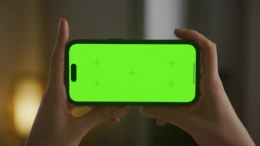 Krom anahtarı olan yatay akıllı telefon, telefon ekranına doğru yavaşça yakınlaştır. Beyaz kadın elleri kapalı alanda yeşil ekranlı akıllı bir telefon tutuyor.