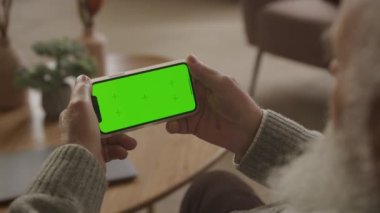 Dairesinde yatay yeşil ekran akıllı telefona bakan yaşlı bir adam, beyaz sakallı yaşlı bir adam omuz manzarası üzerinden krom anahtar akıllı telefonu izliyor.