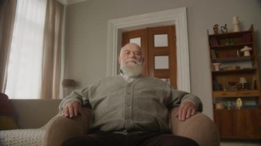 Üst düzey adam kameraya bakıyor, düşük açılı geniş açılı çekim dairede, büyükbaba sandalyede oturuyor ve kameraya uzanıyor.