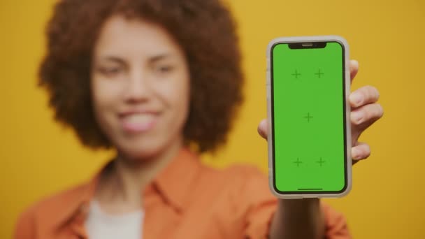 緑の画面のスマートフォンを示す女性は黄色の背景にクローズアップ 女性はクロマキースマートフォンと笑顔を保持 — ストック動画