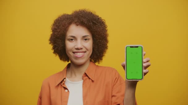在黄色背景下手持绿色荧幕智能手机的女性 现代女性展示垂直彩色按键智能手机 — 图库视频影像