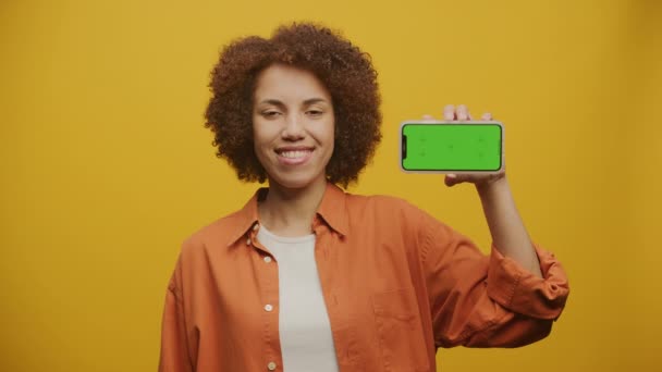 在黄色背景下手持水平绿色屏幕智能手机的妇女 现代女性展示彩色密钥智能手机 — 图库视频影像
