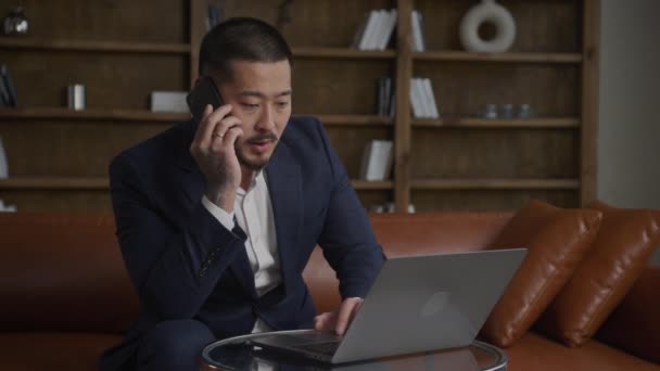 Salesmanの電話 ノートパソコンで働く男 大人入れ墨アジアの男でスーツで仕事の呼び出しとブラウジング上の彼のラップトップでロフトオフィスでスローモーション — ストック動画
