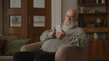 Büyükbaba Smartphone 'a Kol Sandalyesinde Gülüyor, Kıdemli adam internette sörf yapıyor ve gülümsüyor.