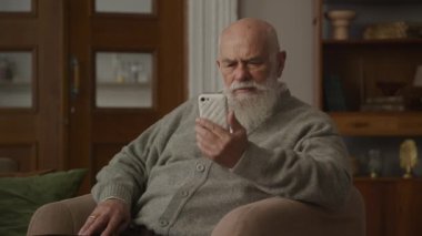 Kollu sandalyedeki akıllı telefondan kötü haberleri okuyan düşünceli yaşlı adam, üzgün büyükbaba baskı altında.