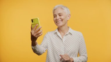 Sarı Arkaplan 'da Olgun Kadın Selamlaması Akıllı Telefon Kullanıyor