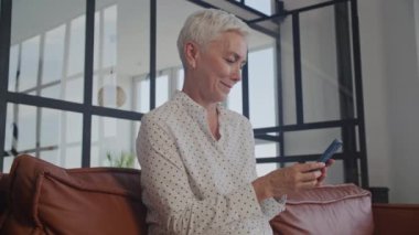 Orta yaşlı kadın akıllı telefonu kapalı mekanda kullanıyor, akıllı telefonda yazan olgun öğretmenin yörüngesinde.