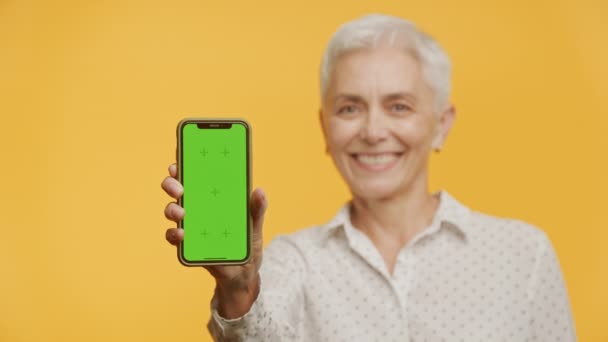 老年妇女在黄色背景下展示绿色屏幕智能手机 — 图库视频影像