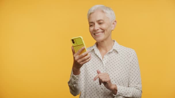 イエローバックグラウンドで電話に応答するシニア女性 スマートフォンを使用して話す高齢女性 — ストック動画