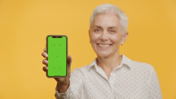 在黄色背景下手持绿色屏幕智能手机的成熟女性 老年女性展示垂直色谱关键智能手机 — 图库视频影像