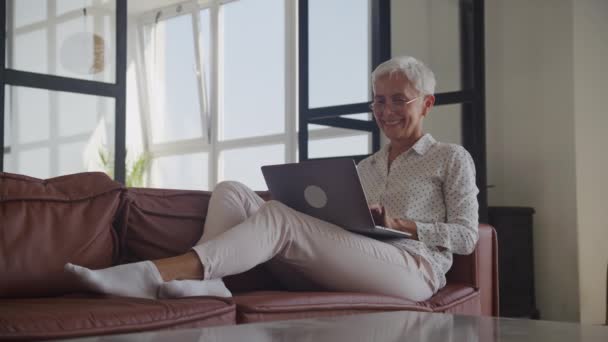 在公寓的沙发上使用笔记本电脑的快乐老年妇女 — 图库视频影像