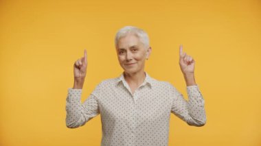 Kısa gri saçlı yaşlı bir kadın iki eliyle işaret ediyor, gülümsüyor ve kameraya bakıyor, sarı arka planda benekli bir bluz giyiyor.