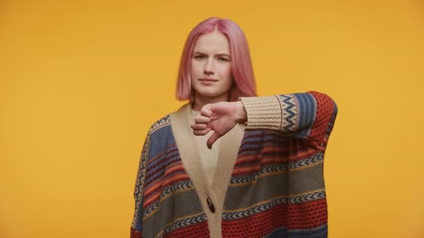 黄色の背景に隔離された多色のセーターを身に着けている親指の下のジェスチャーで不承認を示すピンクの髪の懐疑的なティーン — ストック動画