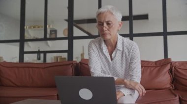 Üst düzey kadın dizüstü bilgisayara odaklandı. Çağdaş bir evde dizüstü bilgisayarla görevlere odaklanan, gözlüklü, yaşlı ve dikkatli bir kadın.