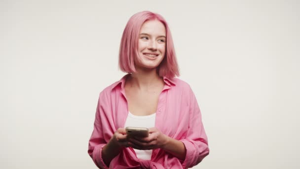 スマートフォンに携わる鮮やかなピンクの髪を持つ笑顔の若い女性 ソーシャルメディアを楽しんだり 白い背景にメッセージを送ったり — ストック動画