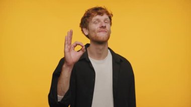 Kıvırcık saçlı genç adam iyi bir jest yapıyor, gözleri kapalı ve kafası hafifçe eğilmiş, içerik bir ifadeyle, hepsi güneşli sarı bir arkaplana karşı.