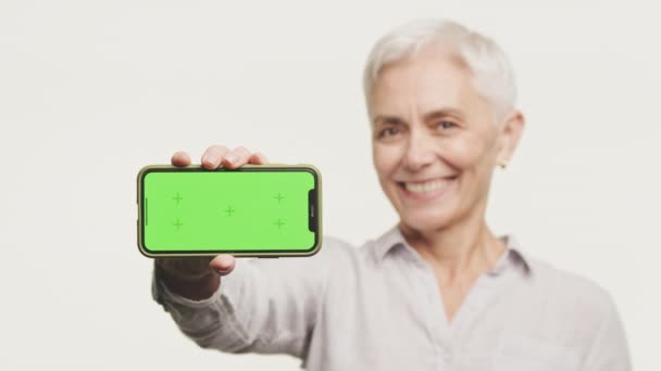 一位年长的女士笑着展示了一款带有绿色屏幕的智能手机 并准备好了定制内容 — 图库视频影像