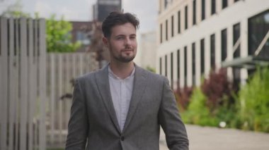 Şık gri takım elbiseli, güler yüzlü genç adam yemyeşil bir şehir bahçesinde güvenle dikiliyor.