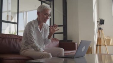 Gözlüklü, gülümseyen yaşlı bir kadın video görüşmesi için dizüstü bilgisayar kullanıyor, sohbet ederken el kol hareketi yapıyor.