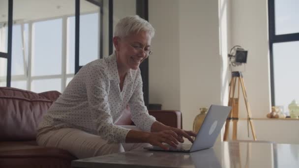 在灯光明亮的现代化房间里 一位戴着眼镜在笔记本电脑上打字的快乐的老年妇女 — 图库视频影像
