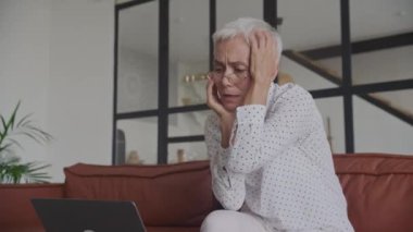 Zor durumdaki yaşlı bir iş kadını, evde dizüstü bilgisayarla çalışırken başını tutuyor.