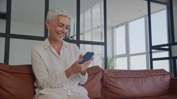 白髪のシニア女性がスマートフォンを見ながら笑い 明るい部屋に革ソファに座る — ストック動画
