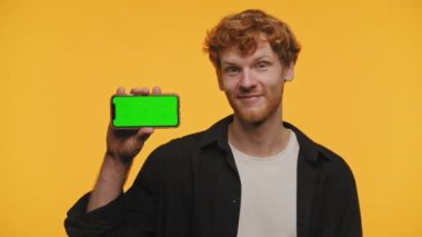 Yeşil ekranlı neşeli adam telefonu tutuyor, sarı arka planda jest yapıyor.