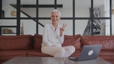 Mutlu, olgun bir kadın barış işareti gösteriyor. Evdeki kanepede dizüstü bilgisayarla oturuyor.