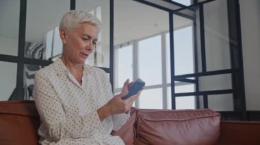 Modern bir ev ortamında deri bir kanepede dinlenirken akıllı telefonunu kullanan kısa beyaz saçlı neşeli bir son sınıf öğrencisi.