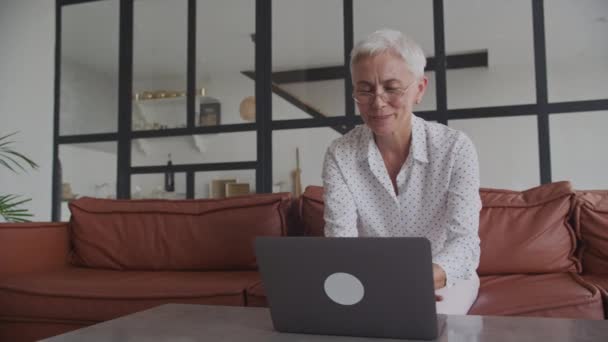 在一间有棕色皮沙发的现代客厅里 一个戴眼镜 留着短发 笑着在笔记本电脑上工作的成熟女人 — 图库视频影像