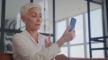 Kısa beyaz saçlı, nişanlı bir kadın akıllı telefonuyla video görüşmesi yapıyor. İyi aydınlatılmış modern oturma odasındaki deri koltukta rahatça oturuyor.