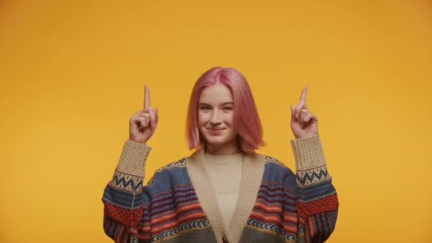 黄色の背景に模様のセーターを着て上向きを指して染められたピンクの髪を持つ若い少女 — ストック動画