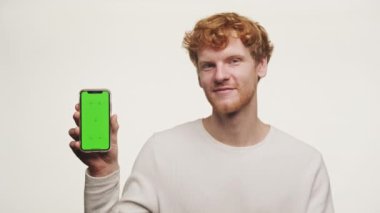 Yeşil ekranlı akıllı bir telefon tutan neşeli kızıl adam, beyaz bir arka planda izole edilmiş, iyi bir jest yapıyor.