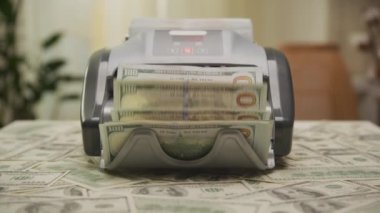 Etrafı dağınık yüz dolarlık banknotlarla çevrili bir masada para sayma makinesi.