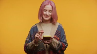 Pembe saçlı neşeli genç bir kadın telefonunda canlı sarı bir arka plana mesaj atıyor.