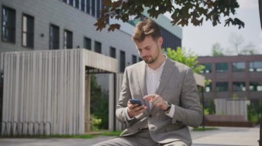 Takım elbiseli profesyonel bir adam şehir parkındaki bankta otururken akıllı telefonuna odaklanmış.