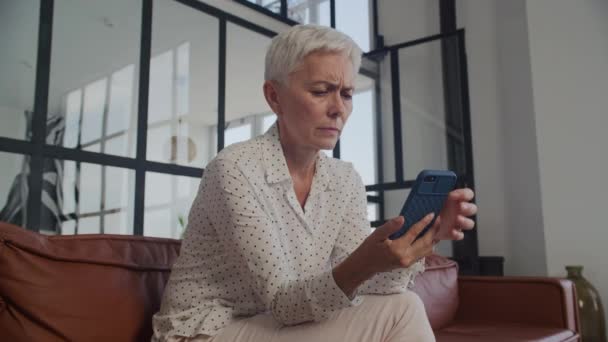 ソファーに座って 心配な表情で彼女の携帯電話を見ている銀髪の苦しみの老婦人 — ストック動画