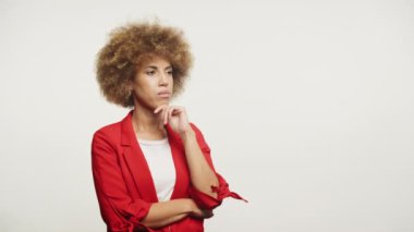Şehvetli afro saçlı, parlak kırmızı ceketli, elleri çenesinde beyaz bir arka planda poz veren düşünceli genç bir kadın.