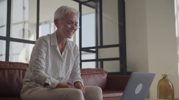 一个快乐而成熟的女人 戴着眼镜 带着笔记本电脑 躺在皮革沙发上 这反映了家里的休闲和连接 — 图库视频影像