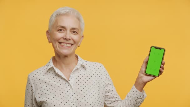 在黄色背景下 微笑的老年女性展示绿色屏幕的手机 适合应用演示 — 图库视频影像