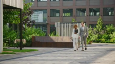 Çağdaş bir şehir bahçesinde bir pano ve dizüstü bilgisayarla yürüyen iş ikilisi