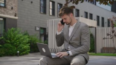 Takım elbiseli genç profesyonel bir adam dizüstü bilgisayar kullanıyor ve şehir bahçesinde otururken telefonla konuşuyor.