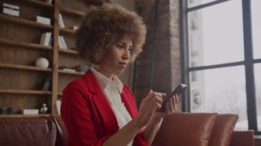 Kırmızı ceketli neşeli iş kadını modern bir çatı katında akıllı telefonuyla sevinçten havalara uçuyor.